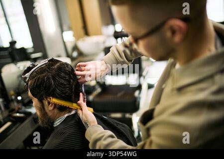 Caucasian young adult man cutting hair of middle aged Caucasian man in barbershop, using scissors and comb, focusing on precise haircutting technique, client sitting in barber chair Stock Photo