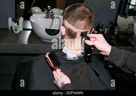Caucasian young adult man sitting in barbershop chair getting haircut, barber using electric hair clipper and brush on back of head, short hair being trimmed, modern salon setting Stock Photo