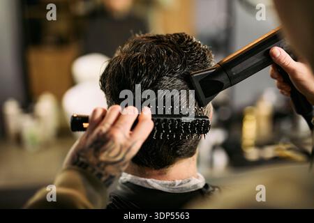 Caucasian young adult man receiving haircut in barbershop, barber with tattooed hand styling hair using round brush and hair dryer, close up of back of head during grooming process Stock Photo