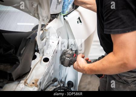 A mechanic uses a power drill to work on the interior metal frame of a white car door inside a body repair shop. Stock Photo
