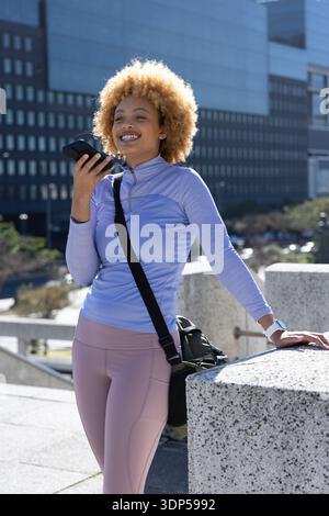 African American woman in activewear holding phone at mouth, crossbody bag and smartwatch on plaza Stock Photo