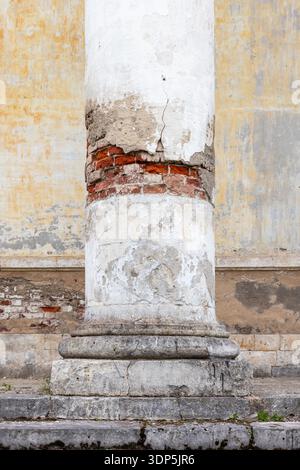 A close-up of a weathered classical column with cracked white plaster revealing exposed red brick, worn stone base and textured facade conveying age, Stock Photo