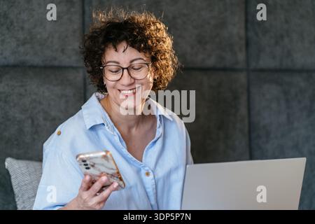 Smiling White woman in her 40s with glasses texting on smartphone while using laptop in bed Stock Photo