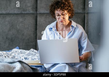 Focused White woman in her 40s with glasses working on laptop in bed at home Stock Photo