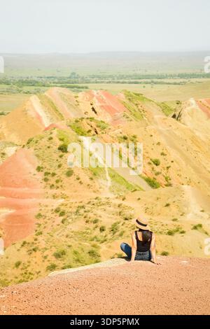 Woman tourist enjoy nature sit relax on colorful vibrant striped hills Mravaltskaro rainbow mountains, Georgia, with unique layered terrain expansive Stock Photo