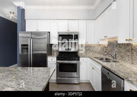 A kitchen with white cabinets, brown and black speckled countertop and backsplash, crown moulding, and grey and blue walls. Stock Photo