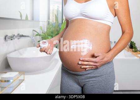 Close-up view of a pregnant woman putting cream of her stretched belly skin in the bathroom Stock Photo