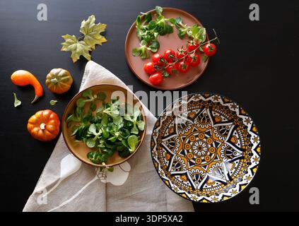 Multicolored ceramic plates and patterned bowl with vegetables and herbs on black background. Food and traditional cuisine. Top view. Stock Photo