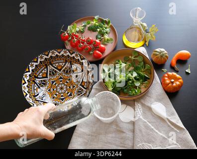 Anonymous person pours water from bottle into glass. Multicolored ceramic plates and patterned bowl with vegetables and herbs on black background Stock Photo