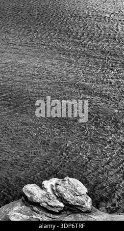 Black and white minimalist coastal scene featuring layered rock formations in the foreground and wind-rippled seawater creating textured patterns acro Stock Photo