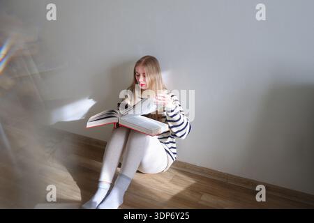 Young woman sitting on wooden floor and reading a large open book at home. Cozy lifestyle, education and self-development concept with copy space Stock Photo