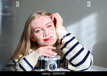 Cheerful blonde woman posing with old fashioned camera at home. Creative profession, personal brand and photography concept. Stock Photo