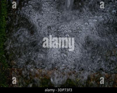 High-angle shot of bubbling water surface creating a complex texture of foam and air bubbles. The dark, moody water contrasts with hints of green moss. Stock Photo