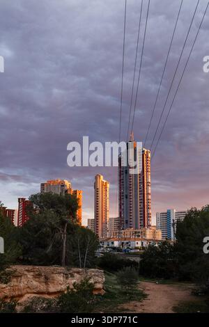 A wide view showing the contrast between the rugged Mediterranean terrain and the towering skyscrapers of Benidorm under a stormy, sunset-tinted sky. Stock Photo