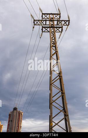 A low-angle shot of a massive steel power transmission tower against a moody, overcast sky, with the iconic skyscrapers of Benidorm visible in the bac Stock Photo