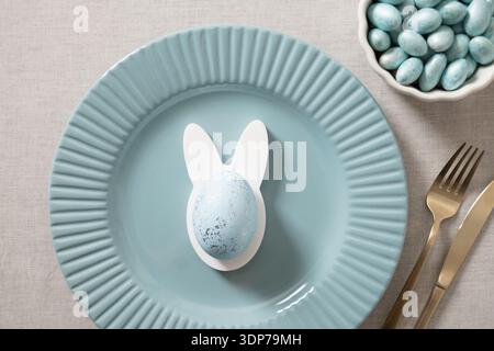Minimal Easter table setting with blue ceramic plate and bunny egg decoration on neutral background, modern festive composition. Stock Photo