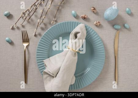 Elegant Easter table setting with blue ceramic plate, linen napkin, golden cutlery and decorative eggs on neutral gray tablecloth. Spring festive dini Stock Photo