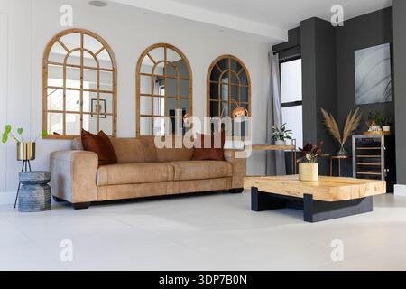 Tan leather sofa is sitting beneath three arched mirrors in bright living room, with rust pillows Stock Photo