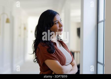 Adult female standing near tall window in bright corridor, looking contemplative with arms crossed Stock Photo