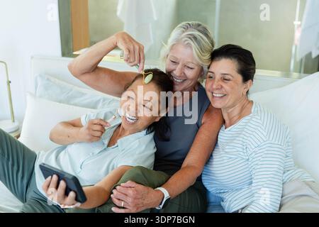 Diverse female friends leaning together on bed, holding smartphone for selfie, using facial rollers Stock Photo