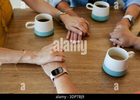 Interlocked adult hands are resting on wooden tabletop near three teal-based coffee mugs Stock Photo