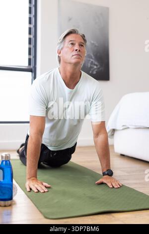 Mature man wearing tee and black shorts practicing backbend on green mat in bedroom near bottle Stock Photo