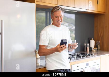Mature man standing in modern kitchen leaning against marble counter holding smartphone and glass Stock Photo