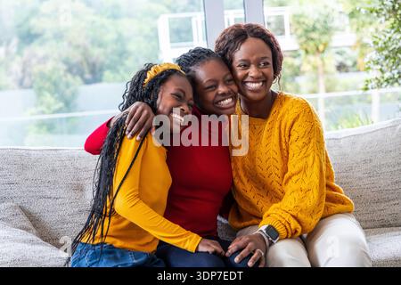 African American mother and teen daughters sitting on light sofa in living room, smiling Stock Photo