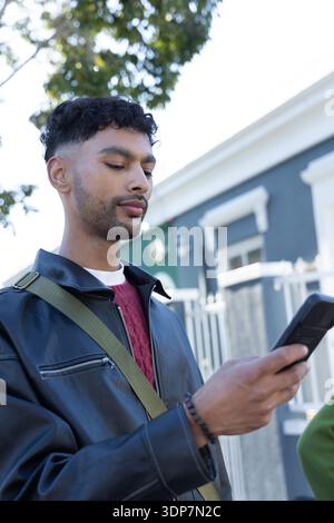 Indian man standing on residential street holding smartphone at chest height wearing leather jacket Stock Photo