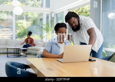 Family father leaning over teenage daughter working on laptop at home while mother using smartphone Stock Photo