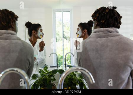 African American couple standing at vanity mirror in bathrobes applying facial mask with brush Stock Photo