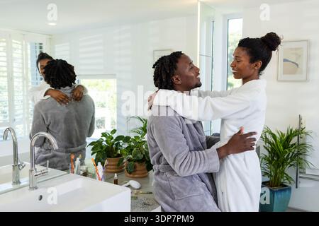 African American couple embracing in bathrobes at vanity, man sitting on countertop, toothbrushes Stock Photo
