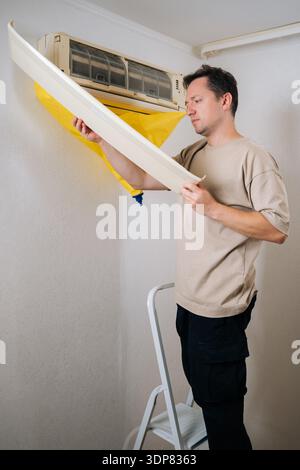 Vertical shot of technician standing on ladder and attaching waterproof bag to air conditioner, preparing for cleaning and maintenance Stock Photo
