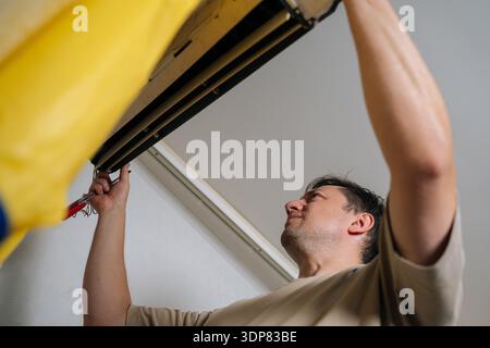 Low-angle view of technician carefully disassembles and repairs wall-mounted air conditioning unit, using screwdriver to adjust internal components Stock Photo