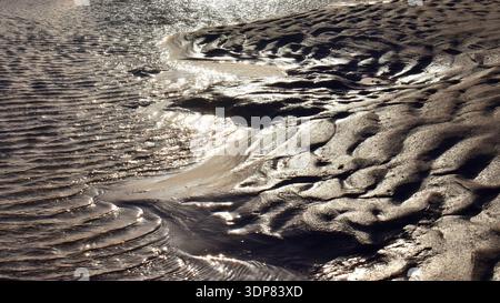 Sunlit water ripples and sand patterns along rocky shore at low tide in widescreen format Stock Photo