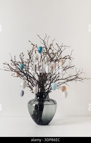 Minimal Easter decoration with branches and hanging pastel eggs in glass vase creating elegant seasonal spring interior. Stock Photo
