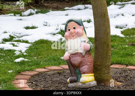 A cheerful garden gnome statue rests beside a tree in a yard, blending whimsy with rustic charm. Snow patches and green grass create seasonal contrast Stock Photo