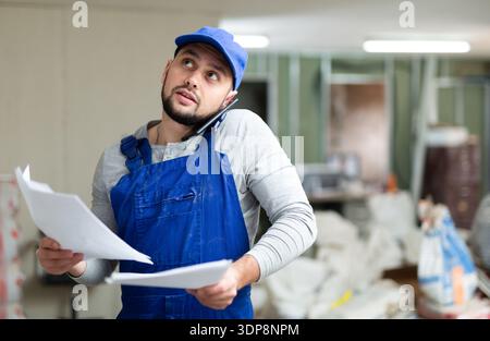Focused bearded contractor in blue overall standing at construction site indoors, discussing work process on phone Stock Photo