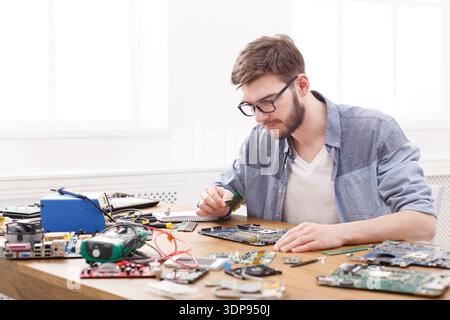 Young Man Works on Repairing Electronic Devices at a Cluttered Table in a Bright Room Stock Photo