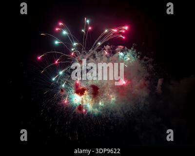 Fireworks: Colorful fireworks exploding in the night sky, with bright red and teal bursts, glowing trails, and drifting smoke against dark background Stock Photo