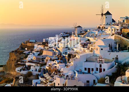 Tourists gather in Oia, Santorini, Greece, to witness the stunning sunset over the Aegean Sea, surrounded by iconic white buildings and windmills. Stock Photo