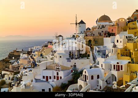 Tourists enjoy the sunset over the iconic white and colorful buildings of Oia, Santorini, Greece, with windmills dotting the cliffside. Stock Photo