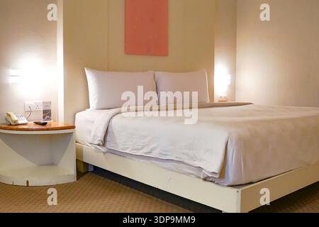 A clean and simple hotel bedroom featuring a neatly made bed with white sheets and pillows, beige walls, and soft carpet flooring, showcasing hospital Stock Photo