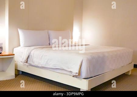 A clean and simple hotel bedroom featuring a neatly made bed with white sheets and pillows, beige walls, and soft carpet flooring, showcasing hospital Stock Photo