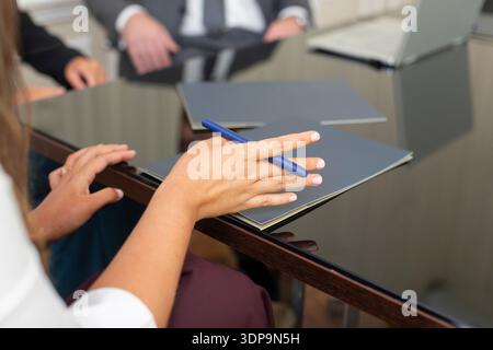 A group of people sits around a table in a meeting room. One person holds a pen and looks at a folder. Papers are on the table along with a laptop. Th Stock Photo