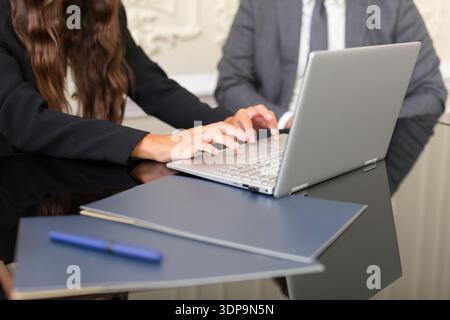 Two individuals are engaged in a business meeting. One person types on a laptop while the other focuses on documents on the table. They are in a profe Stock Photo