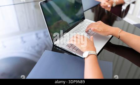A woman is using a laptop at a desk while other individuals are present in the background discussing. The setting is indoors with a bright atmosphere. Stock Photo