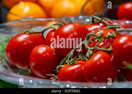 Fresh red tomatoes on the vine in a glass bowl with colorful produce in the background. Stock Photo