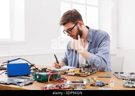 Man Works on Electronics Repair at a Table While Taking Notes in a Bright Room Stock Photo