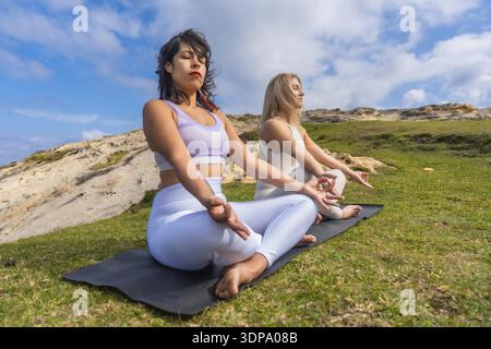 Two young women meditating on yoga mats outdoors, finding peace and mindfulness in nature, practicing spiritual wellness and healthy lifestyle under a Stock Photo